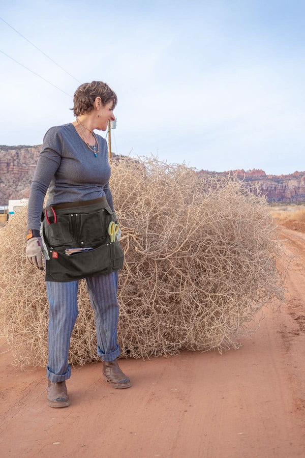 Woman wearing Dovetail Workwear Dovetail Tool Apron in Kodiak Brown with multiple pockets and hammer loops, standing outdoors in desert landscape
