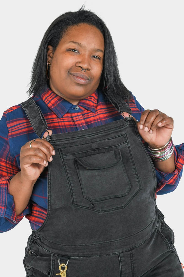 Woman wearing Dovetail Workwear's Freshley Drop Seat Overalls in Black Stretch Thermal Denim with red and blue plaid shirt, holding straps against white background