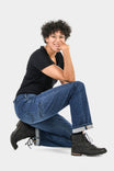 Pride Shop Pant in Indigo Denim Blue by Dovetail Workwear, shown on a woman in a kneeling pose wearing a black shirt and black lace-up boots