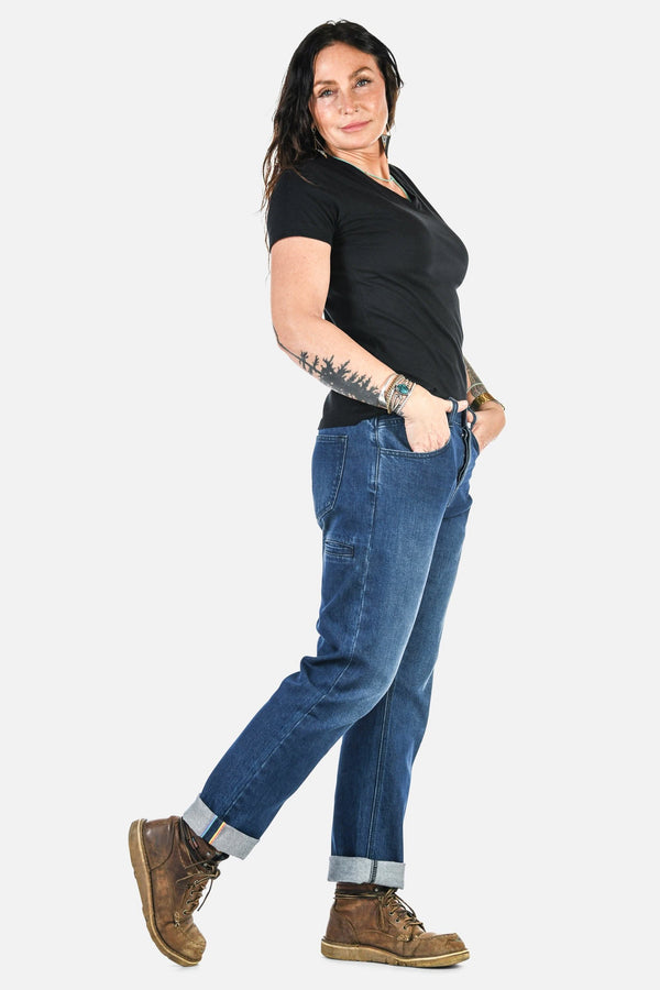 Side view of a woman wearing Dovetail Workwear's Pride Shop Pant in Indigo Denim Blue with rolled cuffs, featuring relaxed leg and multiple pockets