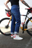 Pride Shop Pant in Indigo Denim Blue by Dovetail Workwear, shown on a woman standing with a colorful bike, featuring rolled cuffs and white sneakers