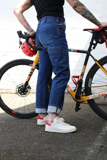Pride Shop Pant in Indigo Denim Blue by Dovetail Workwear, shown on a woman standing with a colorful bike, featuring rolled cuffs and white sneakers