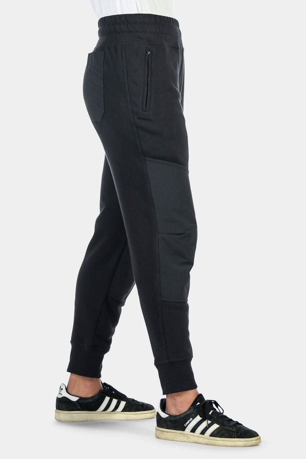 Side view of a woman wearing Dovetail Workwear's black Flagger Fleece Jogger Work Pant, showing reinforced knees, tapered leg, and elastic cuff with black and white sneakers