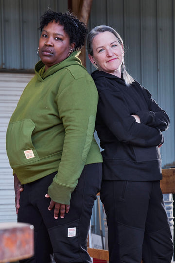 Two women modeling Dovetail Workwear's Flagger Fleece Hoodie in Moss Green, showcasing the reinforced front pocket and thumbhole cuffs from a front-facing angle