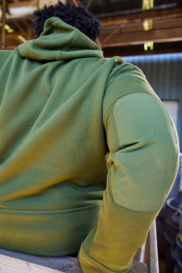 Back view of a woman wearing the Flagger Fleece Hoodie in Moss Green, highlighting the reinforced elbow patches and structured nylon overlays