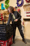 Woman wearing the black M'fon Jacket from Dovetail Workwear in a tool-filled workshop, demonstrating its sleek design and practical features