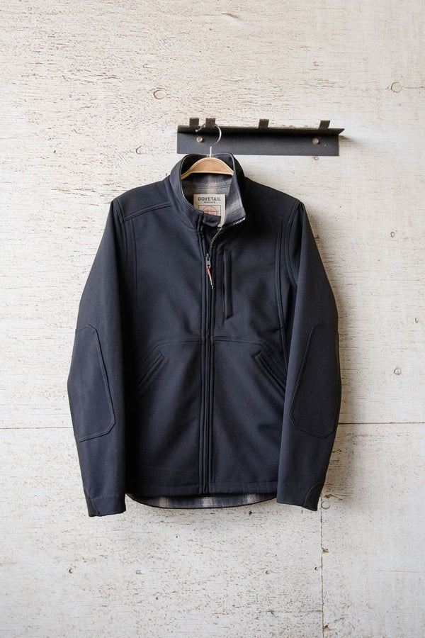Black M'fon Jacket from Dovetail Workwear displayed on wooden wall hanger, showcasing water-resistant softshell exterior, high collar, and deep front pockets
