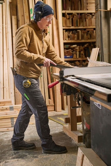 Woman wearing Dovetail Workwear D-Fender Pant in Cement Grey operates a table saw in a woodworking shop, demonstrating the durable work pants' reinforced construction and straight leg design
