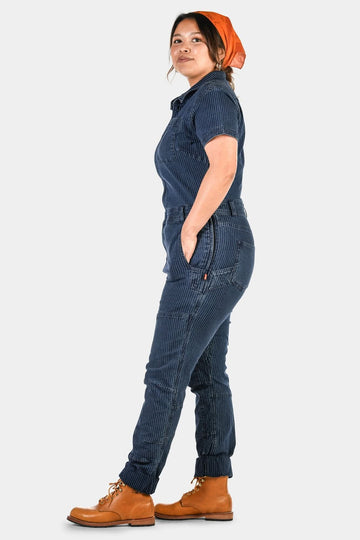 Side view of a woman wearing Dovetail Workwear's Hadley Short Sleeve Coveralls in Dark Blue Wabash Stripe with reinforced knees and elastic cuffs, paired with tan work boots and an orange bandana