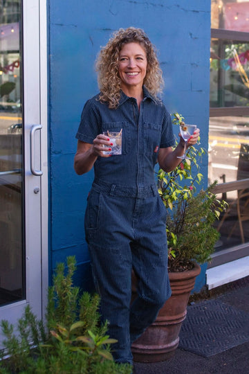 Woman wearing Dovetail Workwear's Hadley Short Sleeve Coveralls in Dark Blue Wabash Stripe, smiling while holding a drink against a blue backdrop with potted plants