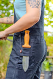 Detail of a woman's tool loop pocket on Dovetail Workwear GO TO™ Stretch Canvas Overalls in Carbon Black, displaying an orange handle and blade against green foliage background