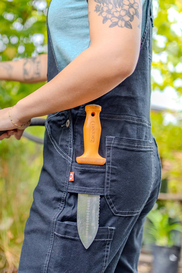 Detail of a woman's tool loop pocket on Dovetail Workwear GO TO™ Stretch Canvas Overalls in Carbon Black, displaying an orange handle and blade against green foliage background