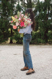 Back view of a woman wearing Dovetail Workwear's Old School Wide Leg Jeans in Indigo Denim with a light blue shirt, holding a large bouquet of pink and yellow flowers