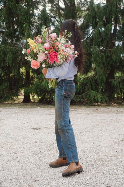 Back view of a woman wearing Dovetail Workwear's Old School Wide Leg Jeans in Indigo Denim with a light blue shirt, holding a large bouquet of pink and yellow flowers