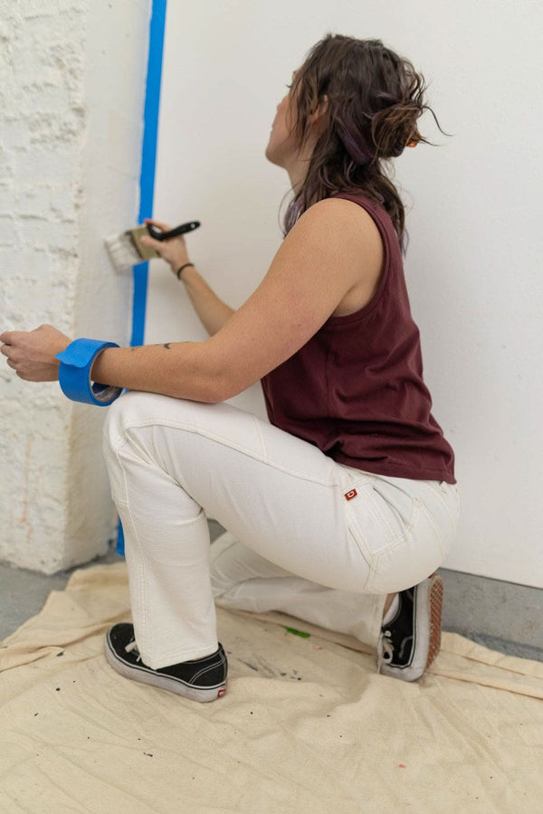 Back view of a woman wearing Dovetail Workwear's GO TO™ Stretch Canvas Work Pant in Painter's White, demonstrating the pant's fit while painting a wall with blue painter's tape