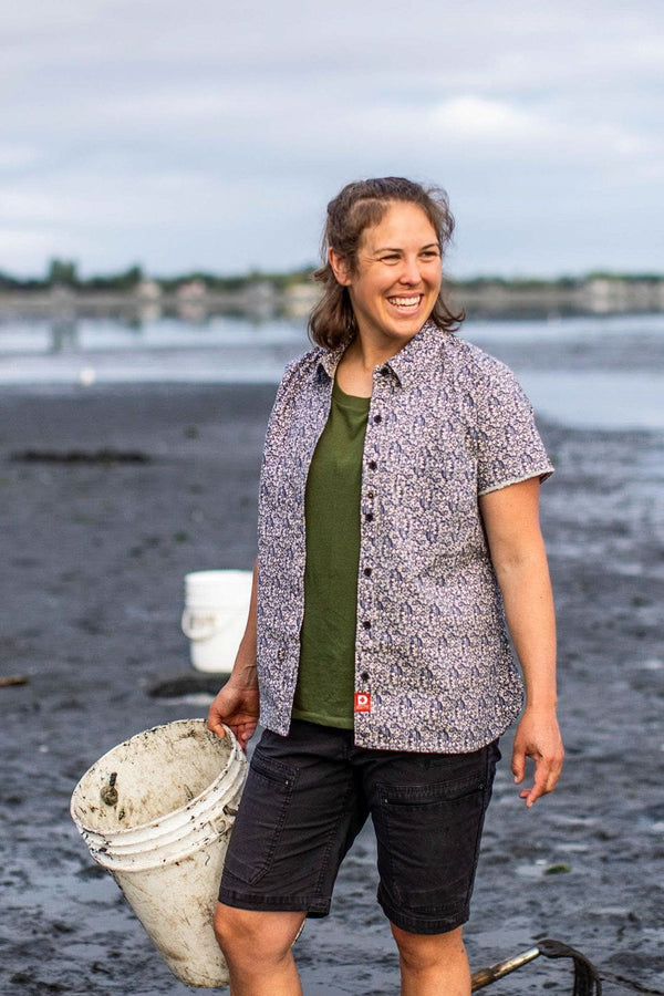 Woman wearing Dovetail Workwear Carver Work Shirt in paprika paisley print with olive tee, holding bucket at beach, smiling at camera