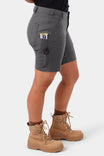 Side view of a woman wearing Dovetail Workwear's GO TO™ Stretch Canvas Short in Cement Grey, featuring visible tool loop and pockets, paired with hiking boots