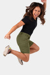 Woman jumping in Dovetail Workwear GO TO™ Stretch Canvas Shorts in Kelp Green, displaying mid-rise fit and 7.5" inseam with visible tool loop and pockets