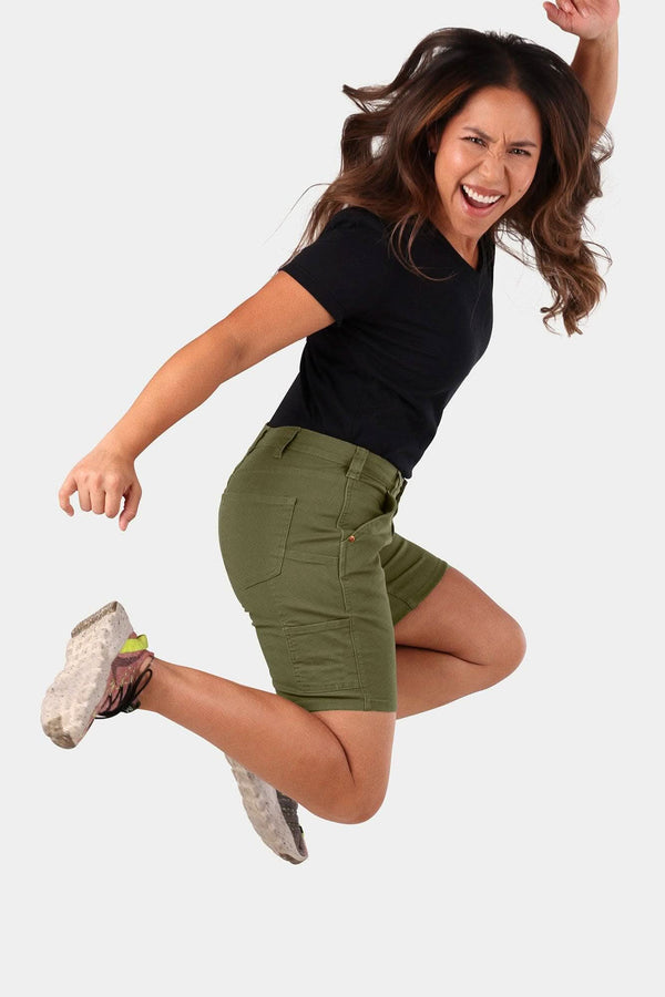 Woman jumping in Dovetail Workwear GO TO™ Stretch Canvas Shorts in Kelp Green, displaying mid-rise fit and 7.5" inseam with visible tool loop and pockets