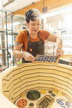 Woman wearing the Kodiak Brown Obana Work Apron by Dovetail Workwear arranging items in one of its pockets while working in a studio setting