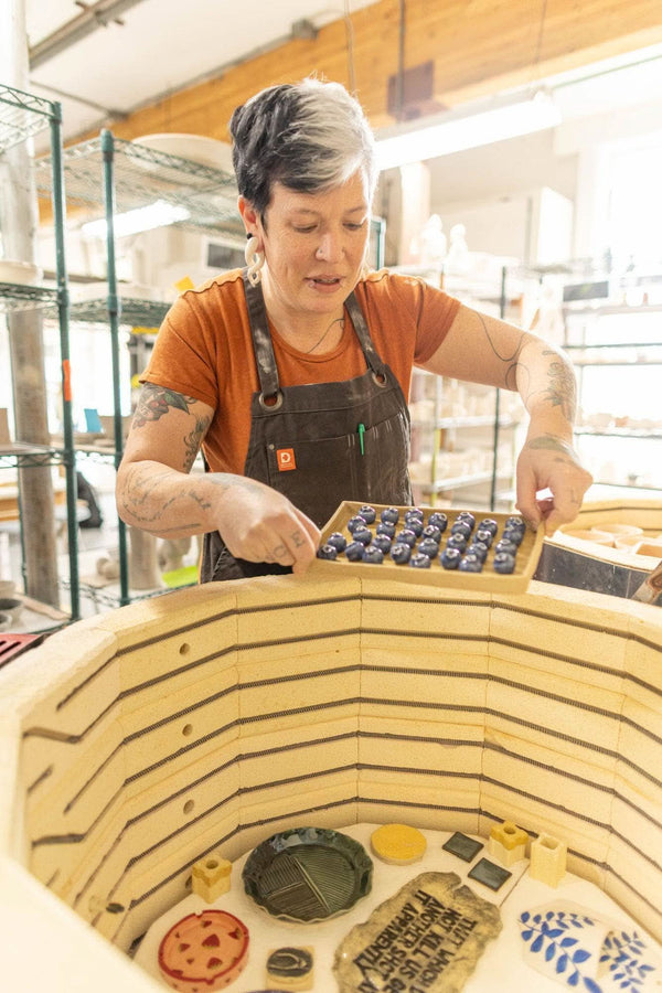 Woman wearing the Kodiak Brown Obana Work Apron by Dovetail Workwear arranging items in one of its pockets while working in a studio setting
