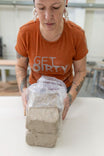 Woman wearing Dovetail Workwear's Get Dirty™ Crew Neck T-Shirt in Paprika (Orange), holding a sand-covered concrete block to demonstrate durability.