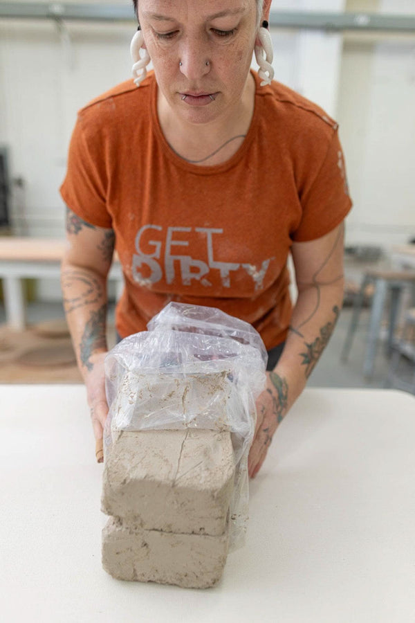Woman wearing Dovetail Workwear's Get Dirty™ Crew Neck T-Shirt in Paprika (Orange), holding a sand-covered concrete block to demonstrate durability.