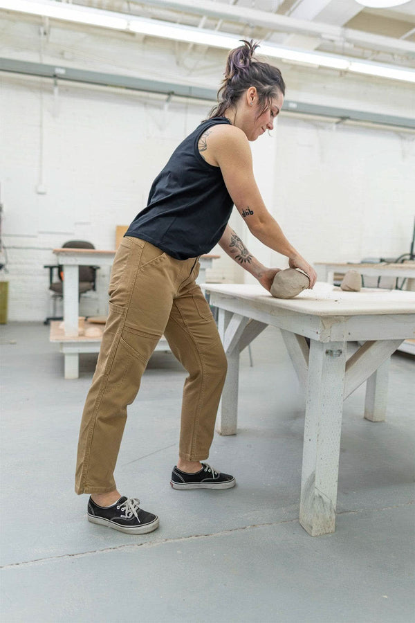 Side view of a woman wearing Dovetail Workwear's Range Crop Pant in Sawdust Brown, demonstrating the relaxed leg and cropped length while working at a table