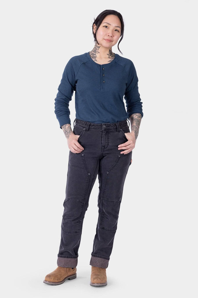 Woman wearing Dovetail Workwear Britt Utility HOTSWAP™ Pant in Black Thermal Denim with navy henley and tan boots, showing the straight leg silhouette and Tough Cuff™ detail