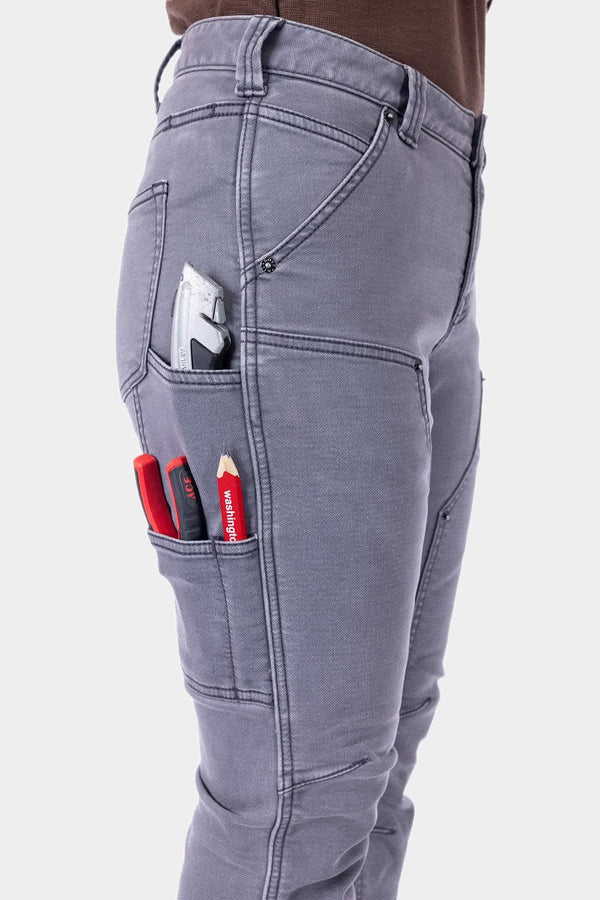 Side view of a woman wearing Dovetail Workwear's Britt Utility HOTSWAP™ Pant in Grey Thermal Denim, displaying multiple cargo pockets with tools and a phone pocket