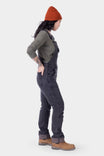 Side view of a woman wearing Dovetail Workwear's Freshley HOTSWAP™ Drop Seat Thermal Denim Overalls in black with a grey thermal shirt and orange beanie
