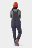 Back view of a woman wearing Dovetail Workwear's Freshley HOTSWAP™ Drop Seat Thermal Denim Overalls in black denim with thermal lining, orange beanie, and hiking boots