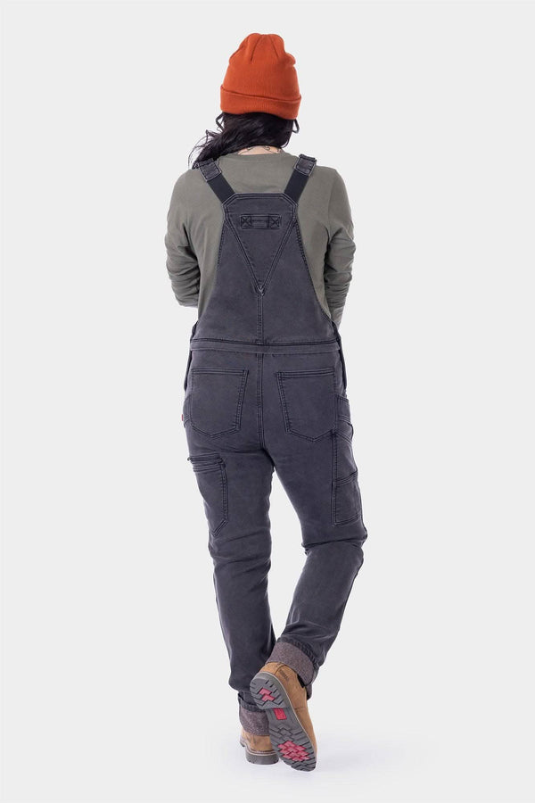 Back view of a woman wearing Dovetail Workwear's Freshley HOTSWAP™ Drop Seat Thermal Denim Overalls in black denim with thermal lining, orange beanie, and hiking boots
