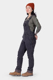 Side view of a woman wearing Dovetail Workwear's Freshley HOTSWAP™ Drop Seat Thermal Denim Overalls in Black with a rust beanie and brown work boots