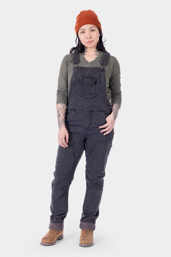 Freshley HOTSWAP™ Drop Seat Thermal Denim Overalls in black by Dovetail Workwear, worn by a woman with long black hair and orange beanie, paired with brown boots