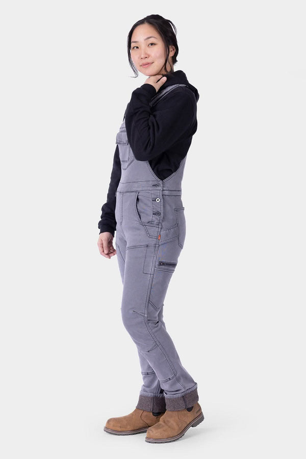 Side view of a woman wearing Dovetail Workwear's Freshley HOTSWAP™ Drop Seat Thermal Denim Overalls in Grey with a black hoodie and brown boots