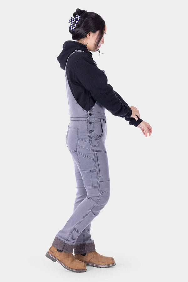 Side view of a woman wearing Dovetail Workwear's Freshley HOTSWAP™ Drop Seat Thermal Denim Overalls in Grey with a black hoodie and tan boots