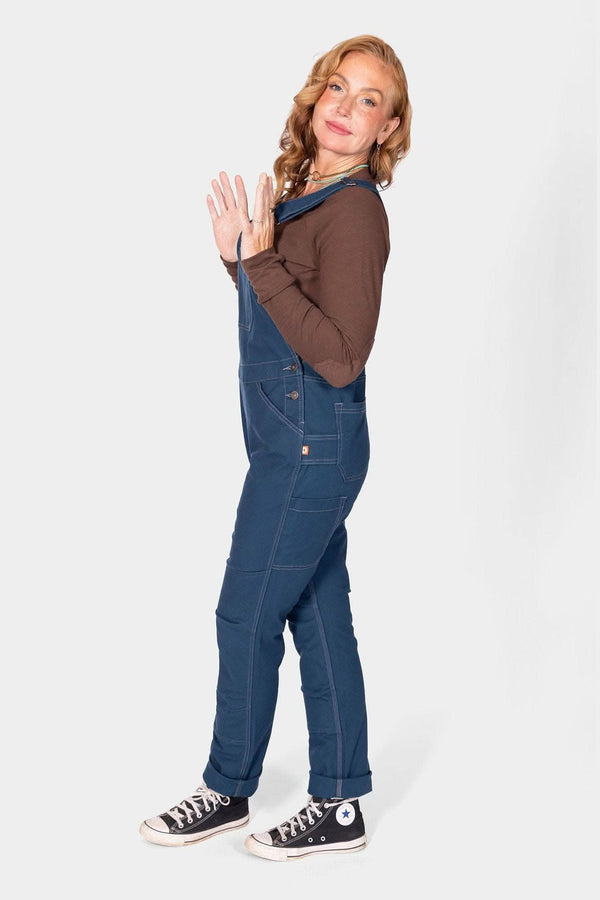 Side view of a woman wearing Dovetail Workwear GO TO™ Stretch Canvas Overalls in Atlantic Blue with brown long-sleeve shirt and black canvas sneakers