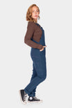 Side view of a woman wearing Dovetail Workwear GO TO™ Stretch Canvas Overalls in Atlantic Blue with a brown long-sleeve shirt and black canvas sneakers