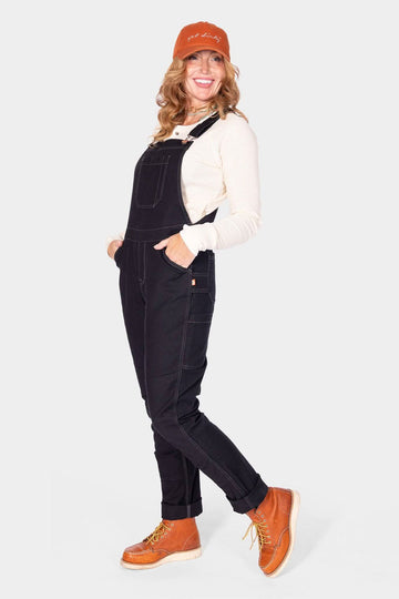 Woman modeling Dovetail Workwear's GO TO™ Stretch Canvas Overalls in Carbon Black, paired with a cream long-sleeve shirt and orange cap
