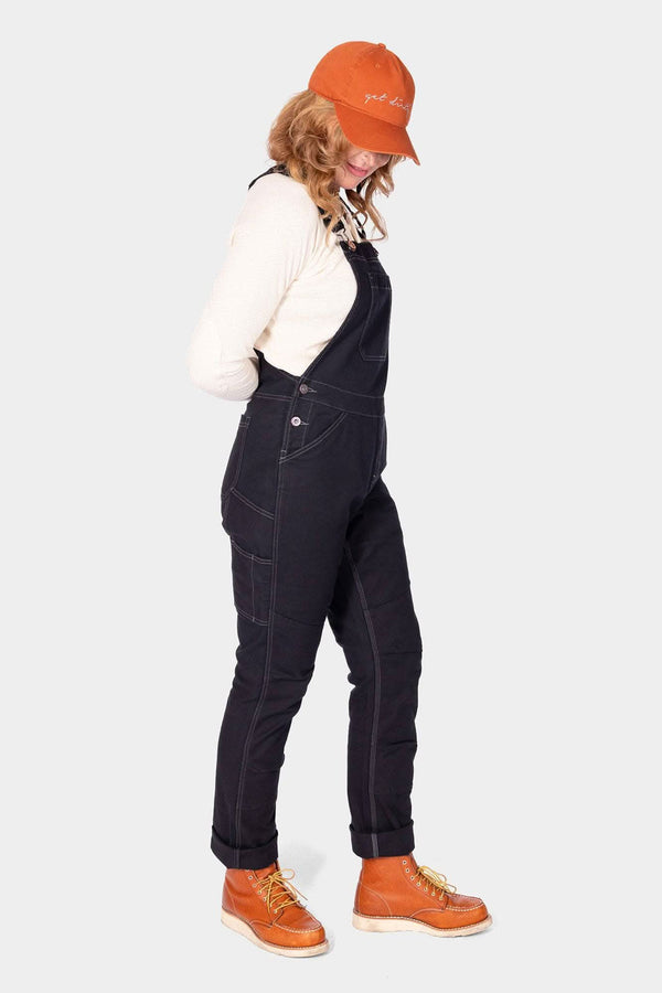 Side view of a woman wearing Dovetail Workwear's GO TO™ Stretch Canvas Overalls in Carbon Black with orange boots and cap
