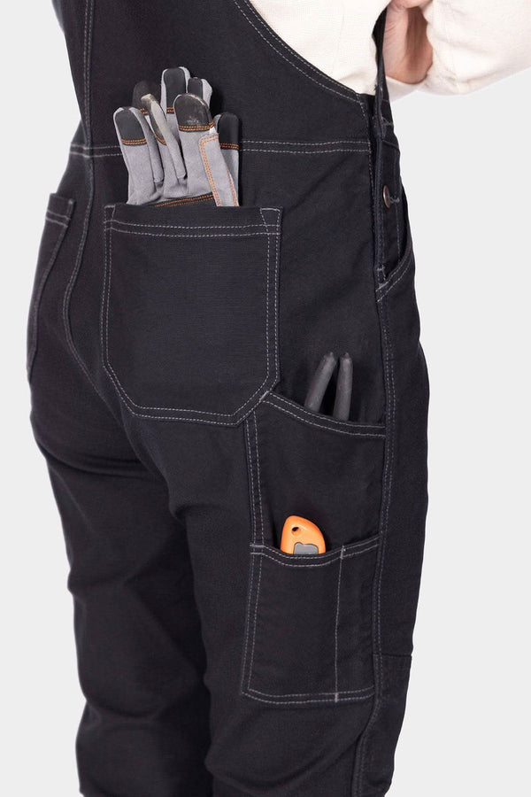 Back view of a woman wearing Dovetail Workwear GO TO™ Stretch Canvas Overalls in Carbon Black, displaying multiple pockets and work gloves stored in the chest pocket