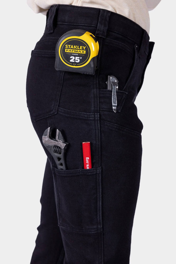 Side view of a woman wearing black Maven X Graphene Denim Pant by Dovetail Workwear, displaying multiple tool pockets and a 25' tape measure on the Hip Slot™