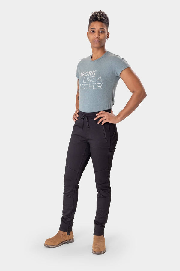 Woman wearing Dovetail Workwear's Christa X DIY Pant in Black Performance Modal with grey "Work Like a Mother" tee and tan boots, displaying nested side pockets and high-rise waistband