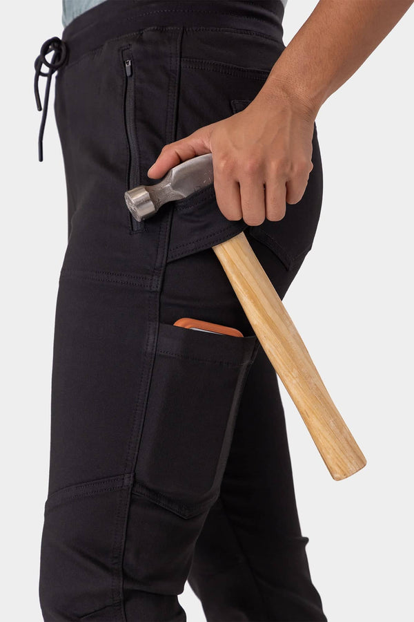 Woman holding a hammer while wearing the Christa X DIY Pant in Black Performance Modal by Dovetail Workwear, showcasing the tool loop and front zip pocket details