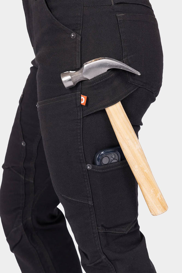 Side view of a woman wearing Dovetail Workwear's Britt Utility Pant in No Fade Black Canvas, displaying the tool loop, Hip Slot™ Zip pocket, and phone pocket while holding a hammer