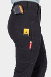 Side view of a woman wearing Dovetail Workwear's Britt Utility Pant in No Fade Black Canvas, displaying nested pockets, tool loop, and a tape measure clipped to the waistband
