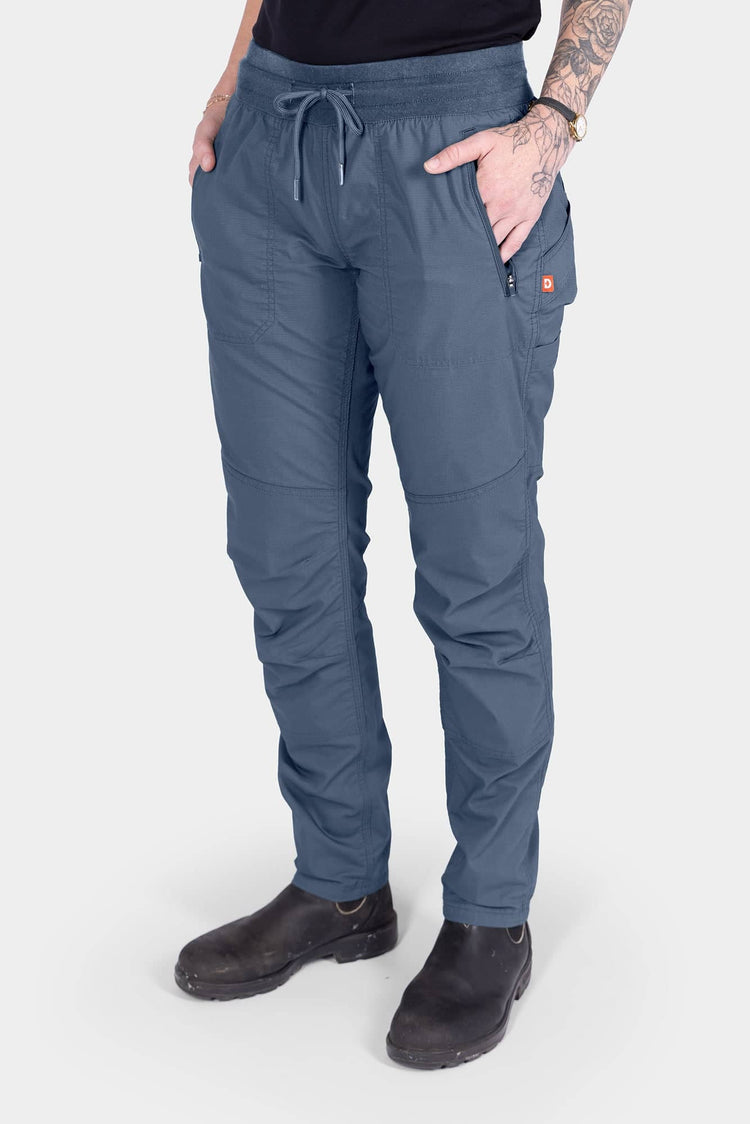 Woman wearing Dovetail Workwear's Christa X COOLSWAP™ Pant in Dovetail Blue, featuring COOLSWAP™ ripstop fabric, front zip pockets, and a high-rise waistband with drawcord