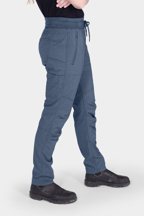 Side view of a woman wearing the Dovetail Workwear Christa X DIY COOLSWAP™ Pant in Dovetail Blue, featuring tool loop, front zip pockets and a high-rise waistband with drawcord