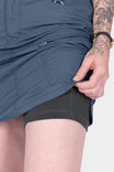 Detail view of a woman's hand opening a bellowed zip cargo pocket on the Dovetail Workwear Compass COOLSWAP™ Cargo Skort in Dovetail Blue, showing functional pocket design